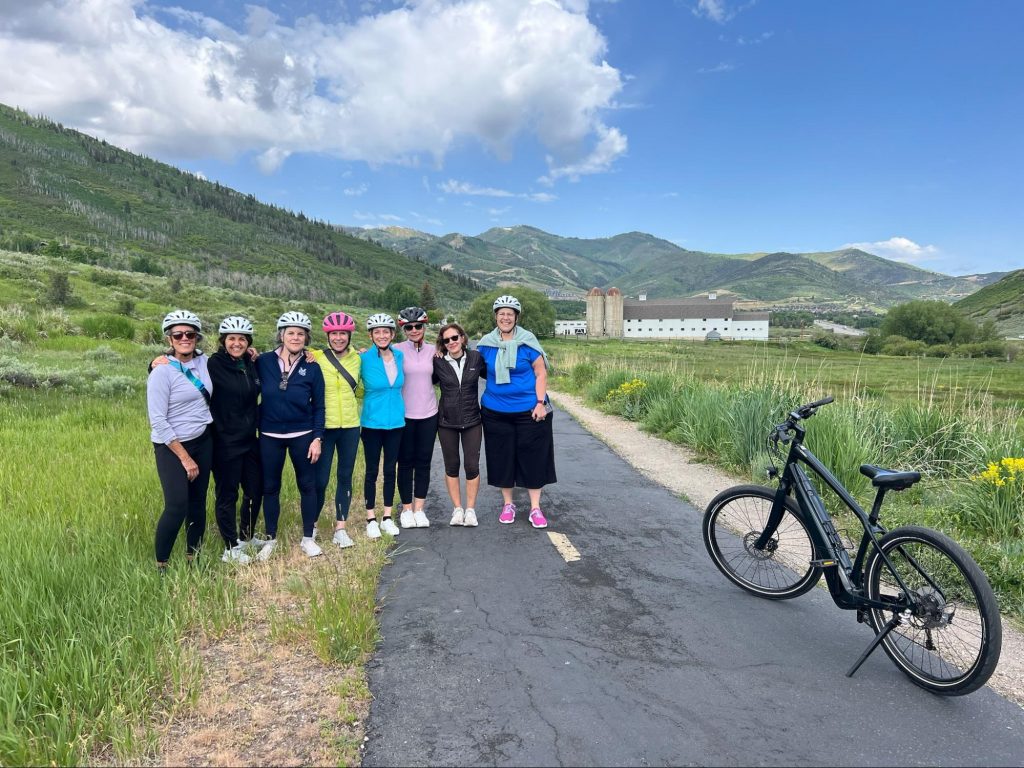 ZizeBikes - Getting Back in the Saddle: A Story of Friendship, Vulnerability, and Finding Your Ride - A group of smiling women wearing bike helmets stands together on a scenic paved trail in Park City, Utah, with green hills and a white barn in the background. An electric bike rests on its kickstand in the foreground. - image1